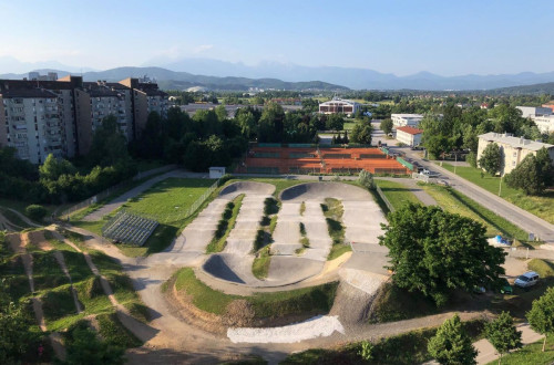 Urbanisticno okolje z BMX poligonom, obkrozeno z zelenimi povrsinami in stanovanjskimi zgradbami, s pogledom na oddaljene gore v ozadju.