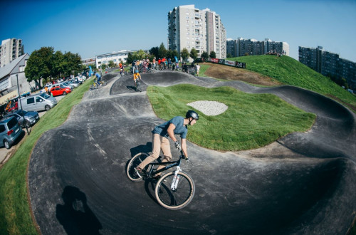 Zivahen prizor na pumptrack poligonu v urbanem okolju, kjer kolesarji razlicnih starosti vozijo po stezi, obdani z zelenimi travnatimi površinami, v ozadju pa so vidni sodobni stanovanjski bloki.