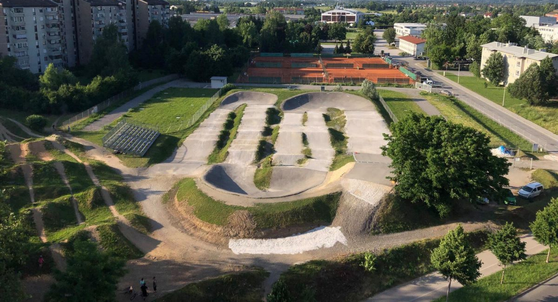 An urban environment with a BMX track, surrounded by green surfaces and residential buildings, with views of distant mountains in the background.