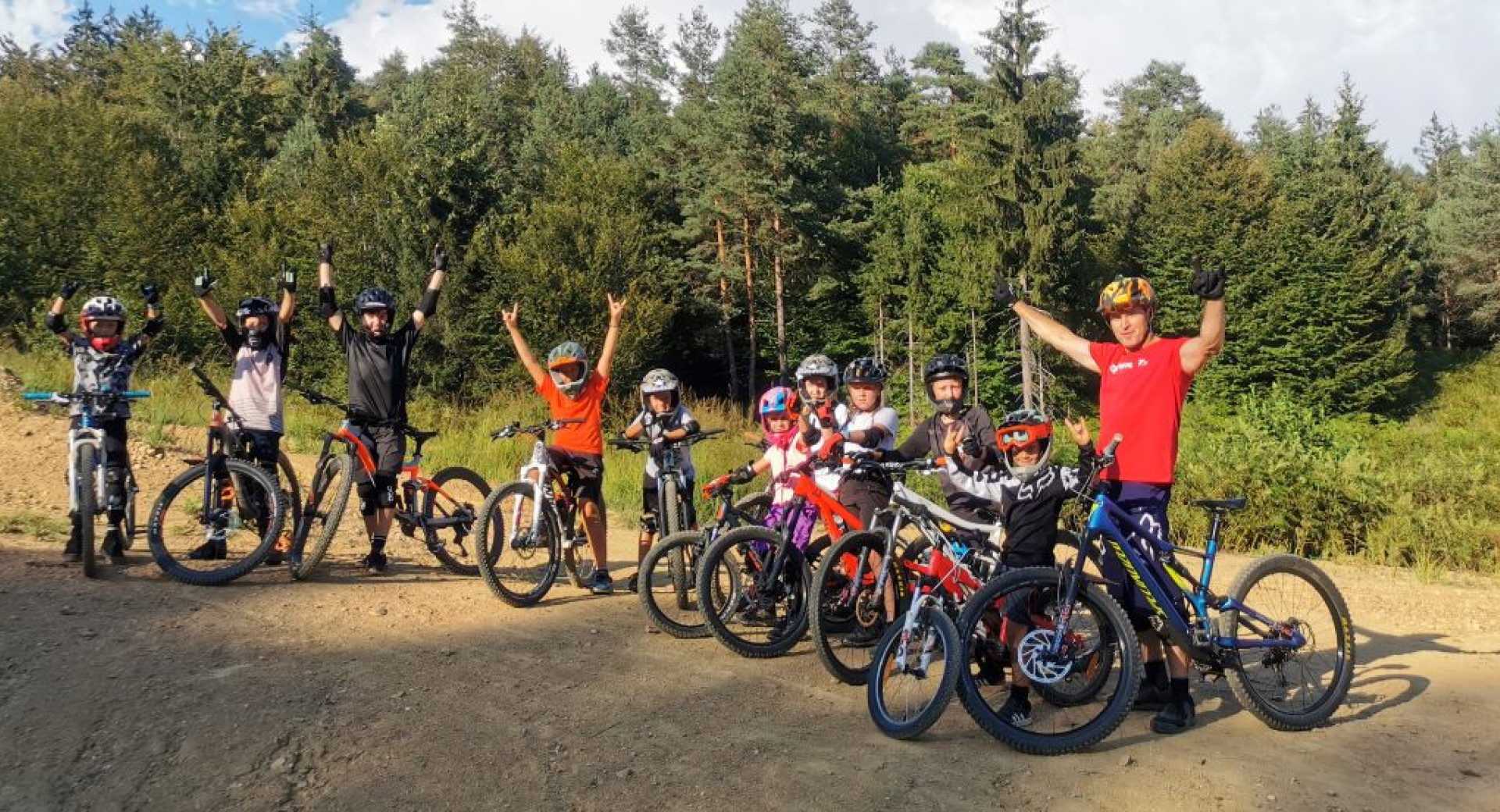 A group of cyclists of various ages stands on a gravel path beside their mountain bikes, with raised arms and smiles, set against the backdrop of a forested area.