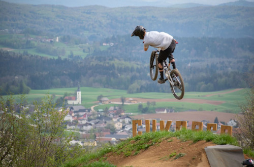 Sportni kolesar na gorskem kolesu izvaja skok na stezi s panoramskim razgledom na podezelsko pokrajino v ozadju, kjer so vidni travniki, cerkev in razprsene hise v dolini.