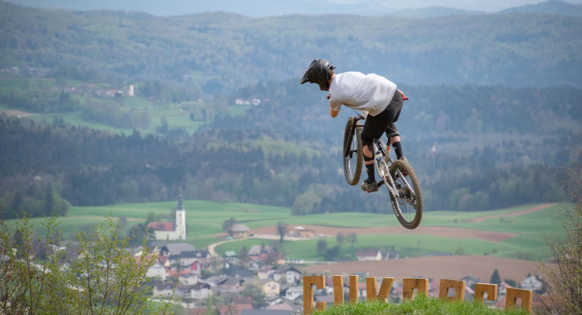 Sportni kolesar na gorskem kolesu izvaja skok na stezi s panoramskim razgledom na podezelsko pokrajino v ozadju, kjer so vidni travniki, cerkev in razprsene hise v dolini.