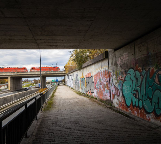 An underpass with a graffiti-covered wall and a view of a railway bridge with a red train passing over the motorway under a cloudy sky.