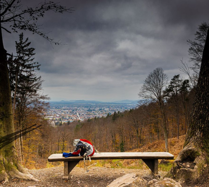 A wooden bench with a rucksack on a forest lookout point offers a view of the city in the distance under a cloudy sky.