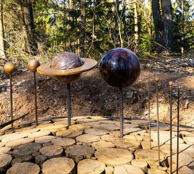 Metal artwork of the planets of the solar system, including Saturn with a ring, set on wooden circular panels in a forest.