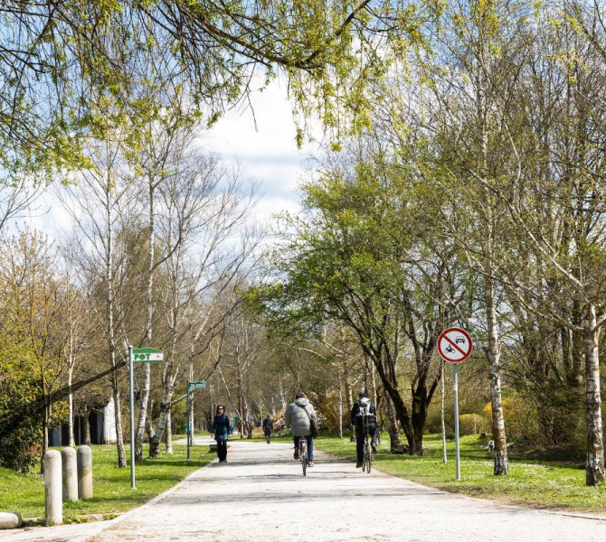 People walk and cycle on the path in the middle of the park, next to a green sign saying 'PST'.