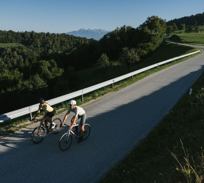 Two cyclists are riding along a winding road, surrounded by green countryside and shaded by trees.