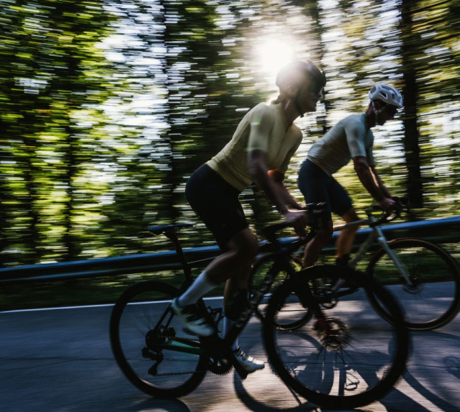 A dynamic image of two cyclists in a forest with sunbeams filtering through the trees.