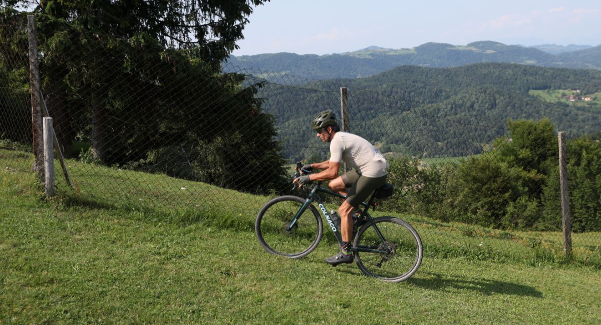 Kolesar v sportni opremi vozi navkreber po travnati poti ob ograji, z razgledom na hribovito pokrajino in gozdove v ozadju.