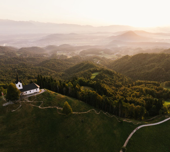 Pogled z visine na cerkvico na vrhu hribcka z ozadjem  gozdnatih hribov.