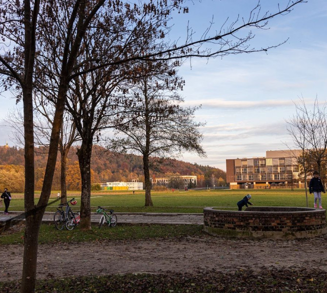 Park z drevesi v ospredju, otroskimi kolesi ob poti, in otroki, ki se igrajo ob nizkem zidanem vodnjaku pod jasnim vecernim nebom.