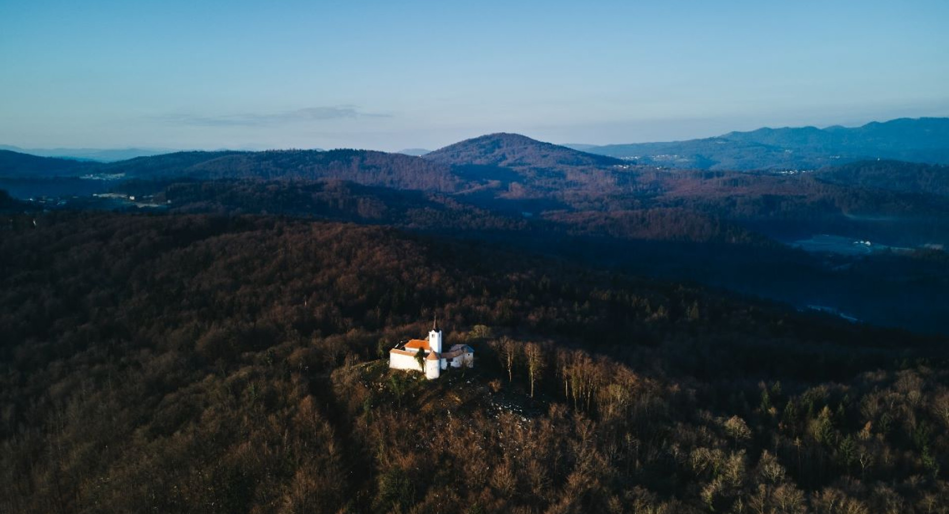 Pogled iz zraka prikazuje majhno cerkev na gozdnatem hribu, obdano z valovitimi hribovji in modrim nebom.