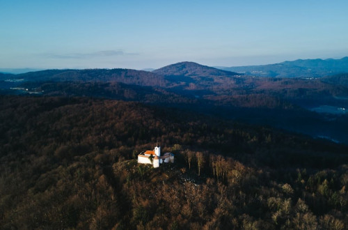 Pogled iz zraka prikazuje majhno cerkev na gozdnatem hribu, obdano z valovitimi hribovji in modrim nebom.