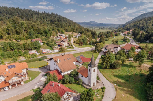 Pogled iz zraka na slikovito vasico, obdano z gozdovi, kjer stoji bela cerkev z zvonikom med hisami.