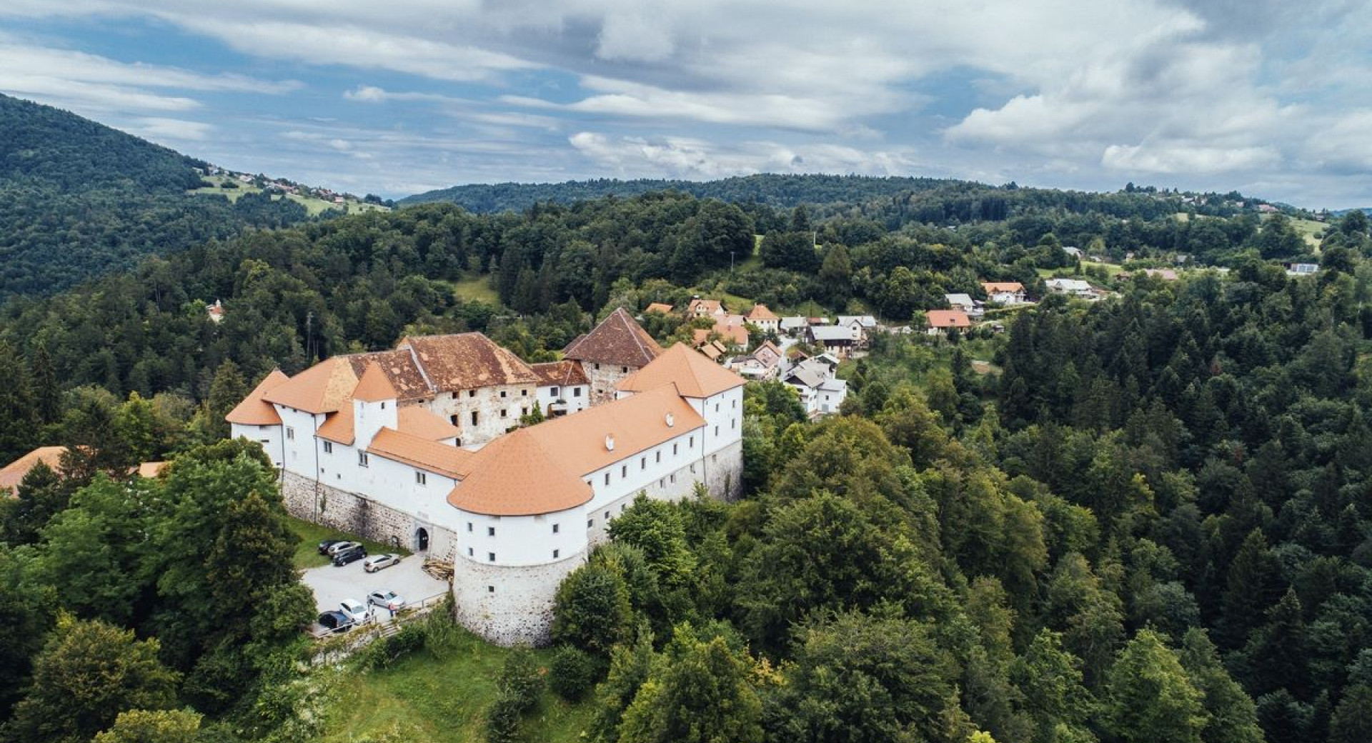 Pogled iz zraka na velik grad z belimi zidovi in rdecimi strehami, obdan z gozdovi in vasjo na hribovitem obmocju.