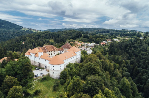 Pogled iz zraka na velik grad z belimi zidovi in rdecimi strehami, obdan z gozdovi in vasjo na hribovitem obmocju.