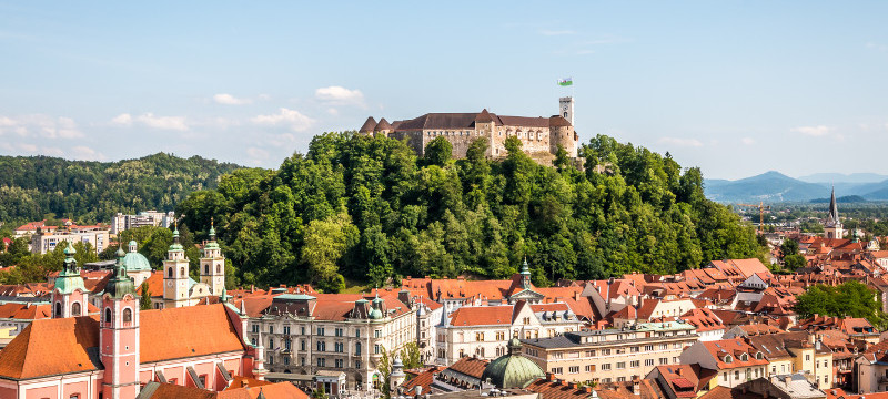 Panoramski pogled na grad, ki stoji na zelenem hribu sredi mesta, obdan z rdecimi strehami mestnih stavb in cerkvenimi zvoniki, pod modrim nebom.