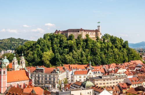 Panoramski pogled na grad, ki stoji na zelenem hribu sredi mesta, obdan z rdecimi strehami mestnih stavb in cerkvenimi zvoniki, pod modrim nebom.