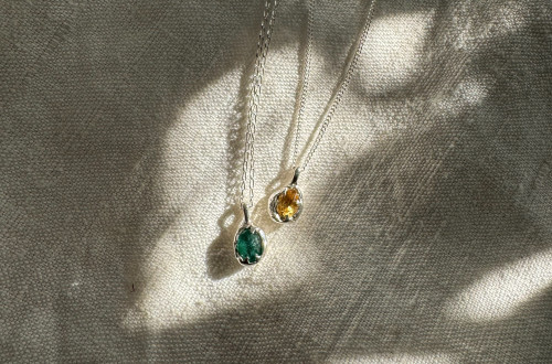 Two silver necklaces with raw green and yellow gemstones, illuminated by soft sunlight on a textured fabric background.