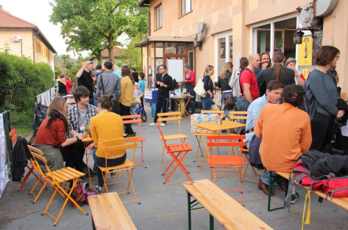 A lively outdoor event with people gathered around small orange tables and chairs, engaging in conversation and enjoying the atmosphere in a neighborhood setting.