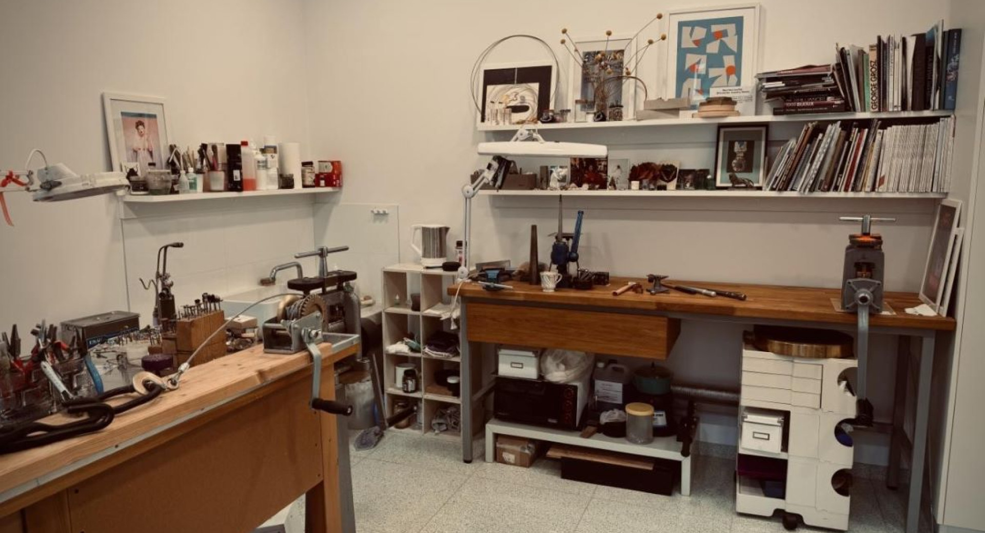 A workspace with tools and materials for jewelry making, featuring a workbench and shelves filled with various items and books.