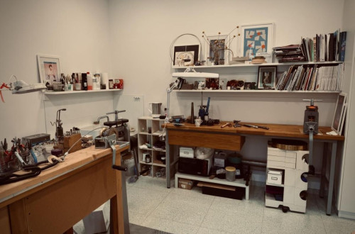 A workspace with tools and materials for jewelry making, featuring a workbench and shelves filled with various items and books.