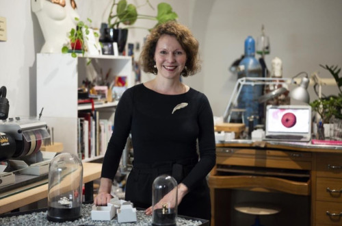 A creative space with the artist standing next to a table, surrounded by materials and tools for creation, with a computer displaying a screen in the background.