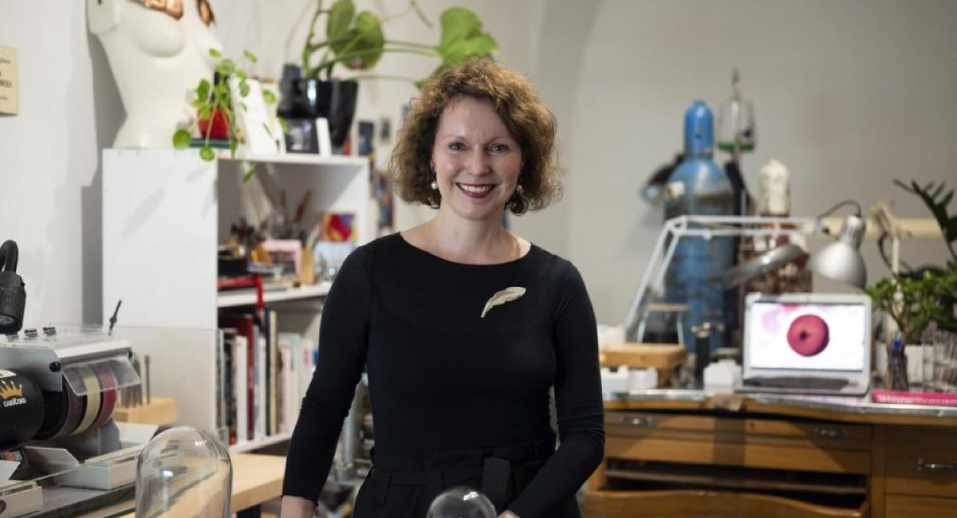 A creative space with the artist standing next to a table, surrounded by materials and tools for creation, with a computer displaying a screen in the background.