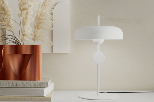 A modern minimalist interior with a white designer lamp, a textured terracotta vase with pampas grass, and a stack of books on a sleek white surface.