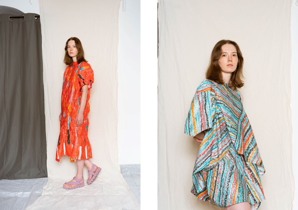 A woman in a loose, colorful striped dress with a draped silhouette, standing against a neutral fabric backdrop.