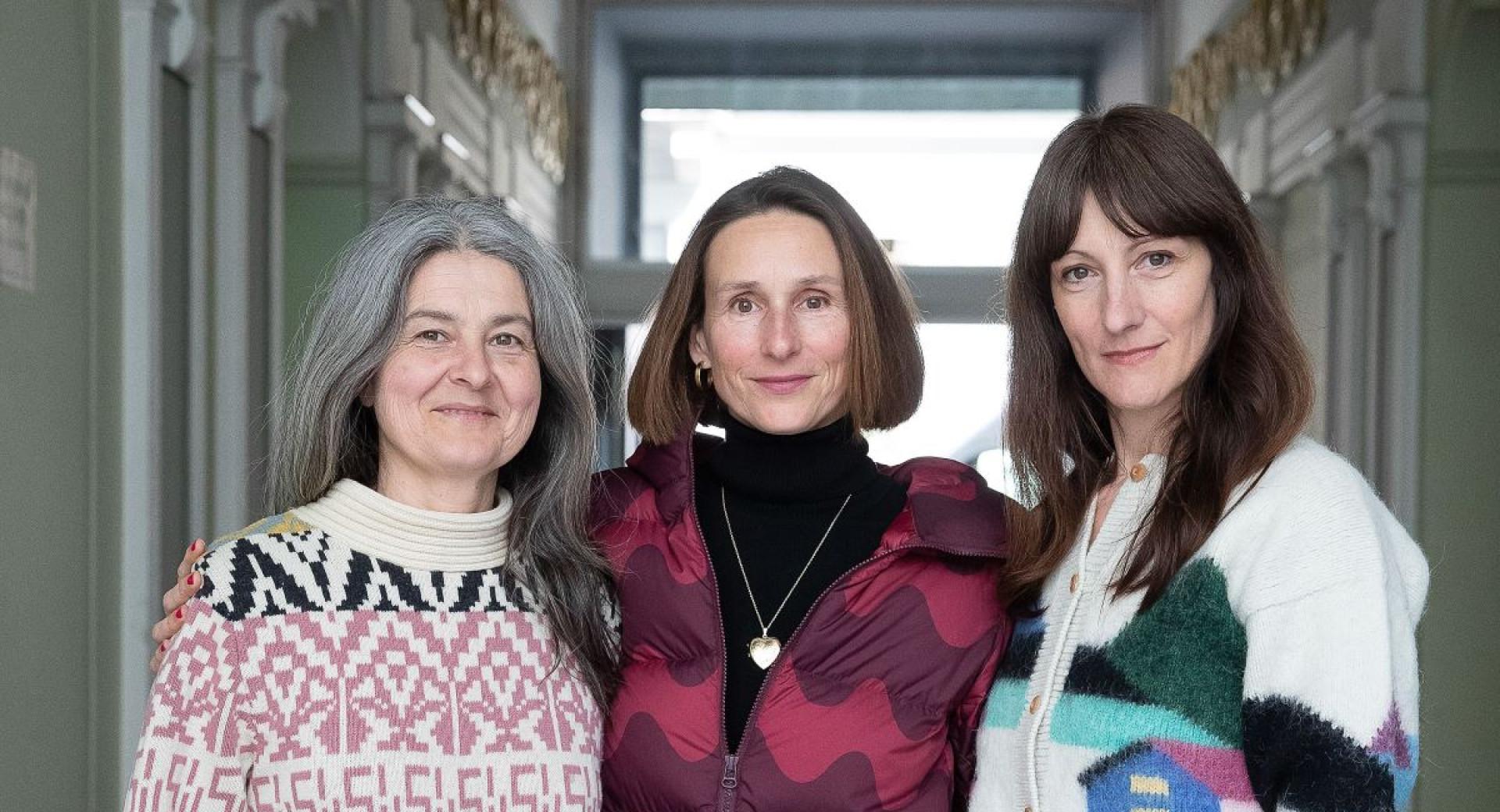 Three women stand together in an elegant hallway, dressed in colorful and patterned knitwear. They are smiling slightly, conveying a sense of warmth and connection.