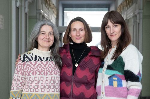 Three women stand together in an elegant hallway, dressed in colorful and patterned knitwear. They are smiling slightly, conveying a sense of warmth and connection.