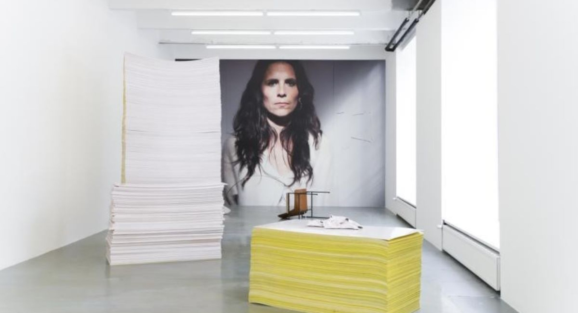 A minimalist exhibition space featuring large stacks of paper and a portrait of a woman in the background.