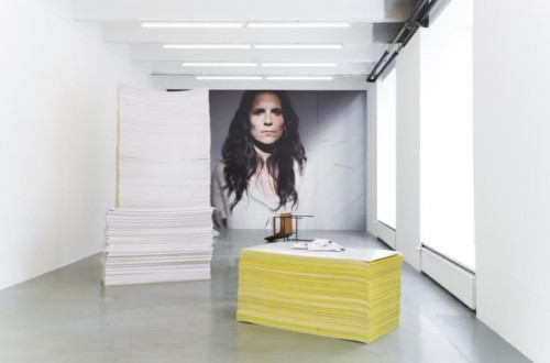 A minimalist exhibition space featuring large stacks of paper and a portrait of a woman in the background.