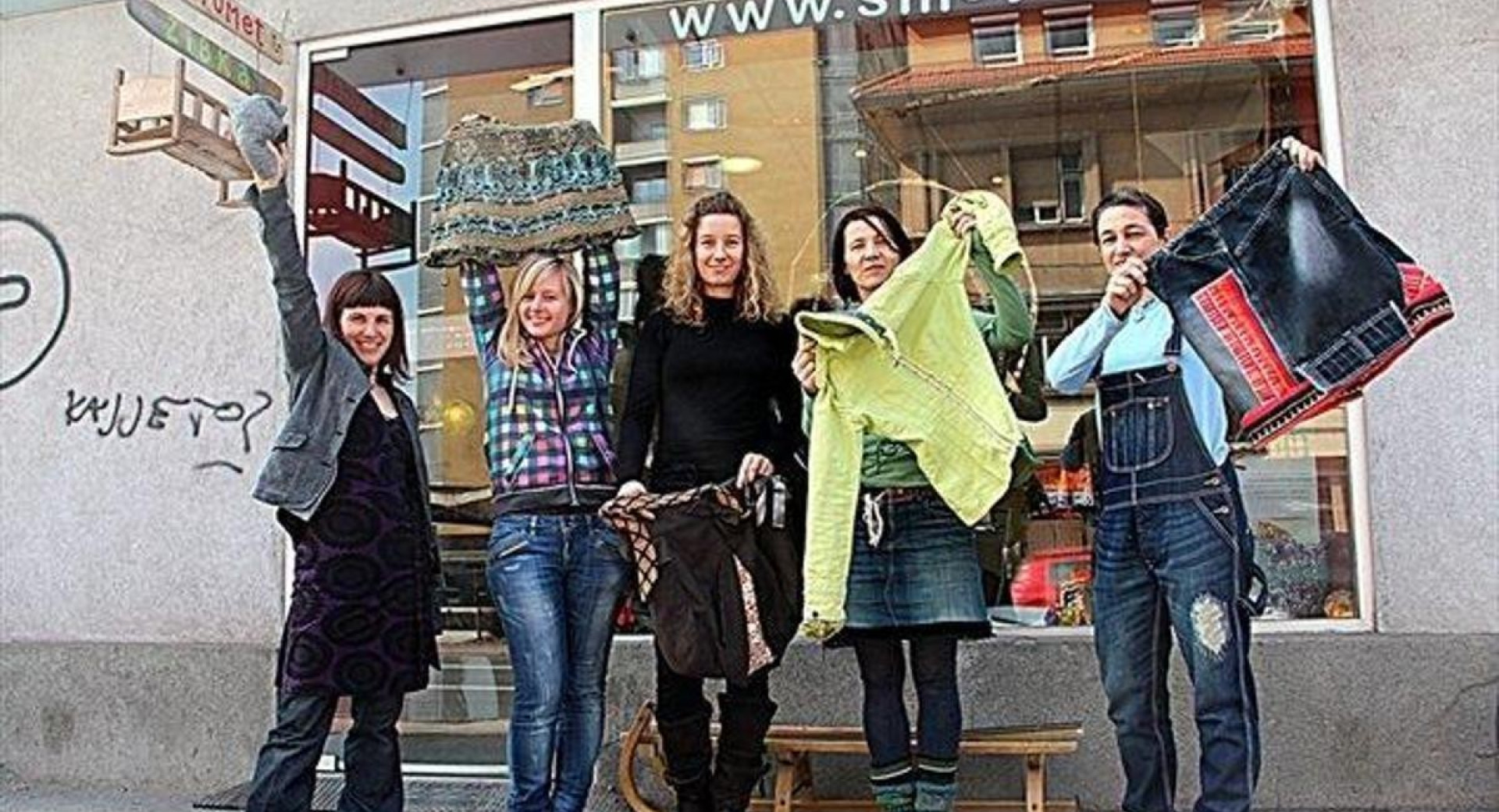 A group of five people standing in front of a shop called "Smetumet," holding up upcycled clothing and accessories.