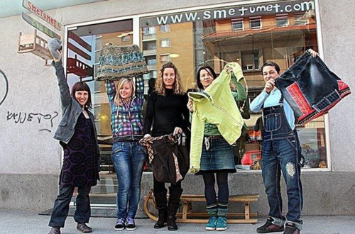 A group of five people standing in front of a shop called "Smetumet," holding up upcycled clothing and accessories.