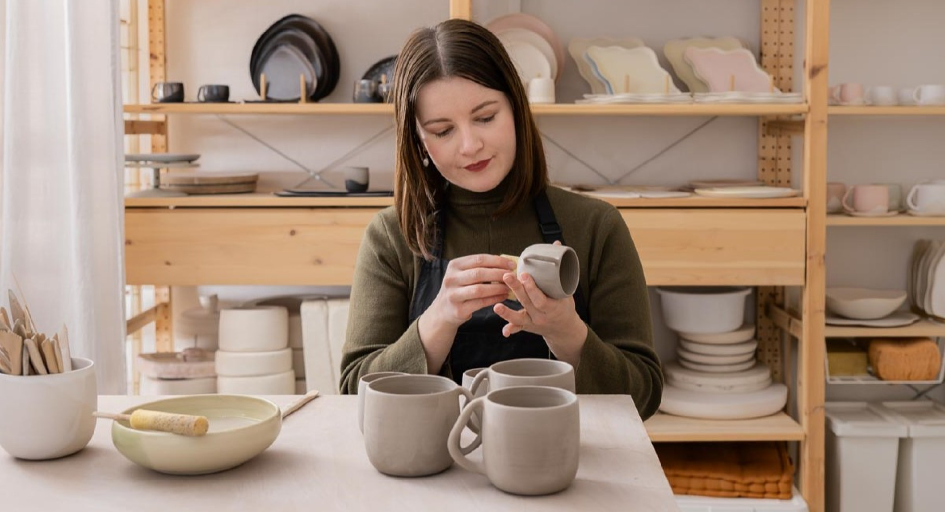 A woman working on a ceramic mug at a table, carefully shaping the details, with shelves of pottery in the background.