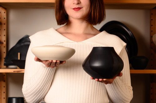 A woman standing in a ceramic studio, holding a black and a white handmade bowl, surrounded by pottery pieces.