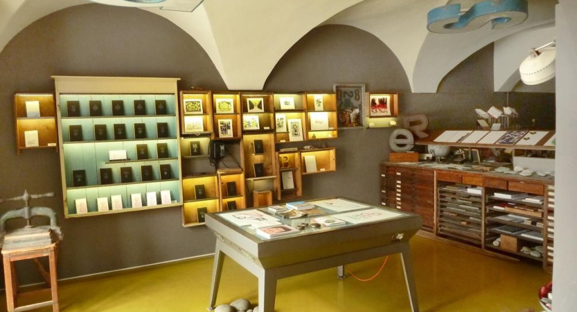 A letterpress studio with arched ceilings, illuminated display cases showcasing printed works, and a central worktable.