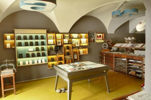 A letterpress studio with arched ceilings, illuminated display cases showcasing printed works, and a central worktable.