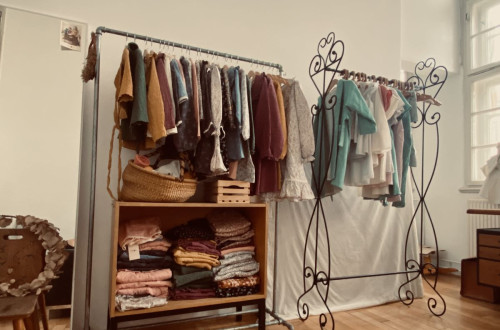 A boutique or showroom displaying handmade, vintage-style clothing on metal racks, with neatly folded textiles on wooden shelves.