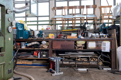 A workshop filled with tools, materials, and plastic baking equipment, with wooden chairs stacked on top of a workbench.