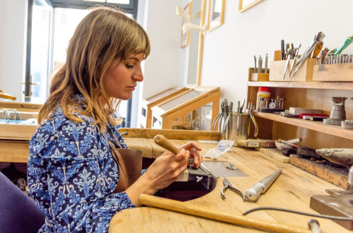 A metal artist working on a jewelry piece at a wooden workbench, surrounded by specialized tools.