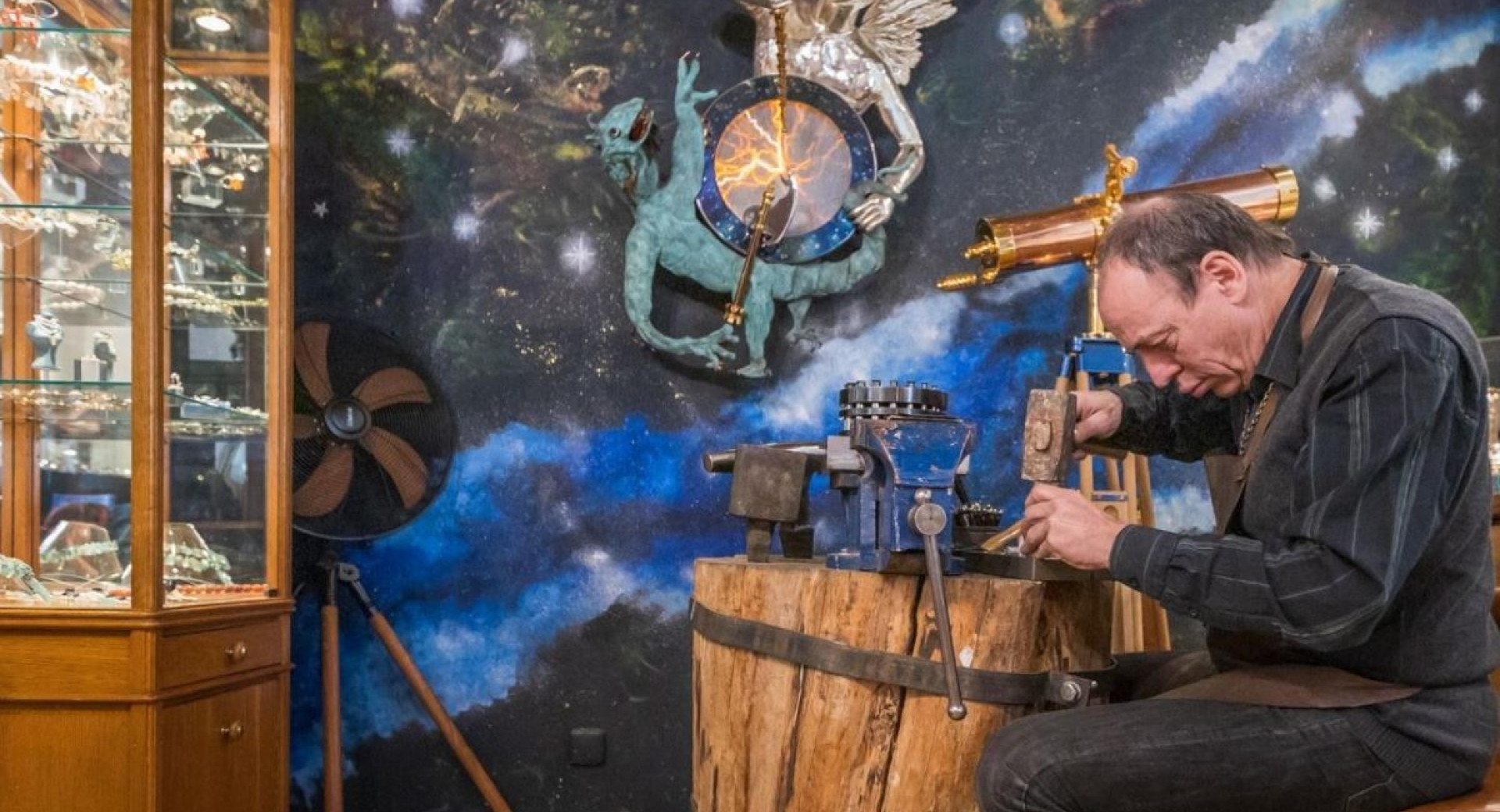 A goldsmith working on a jewelry piece at a wooden workbench, in a shop decorated with celestial and artistic elements.