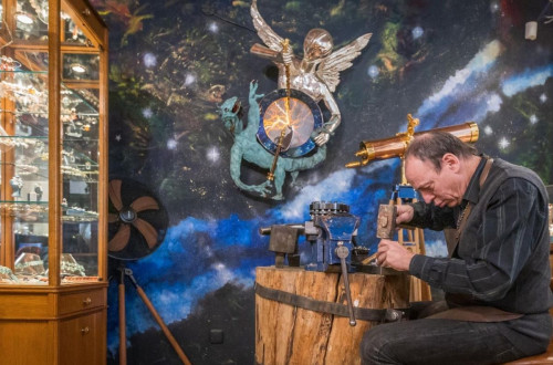 A goldsmith working on a jewelry piece at a wooden workbench, in a shop decorated with celestial and artistic elements.