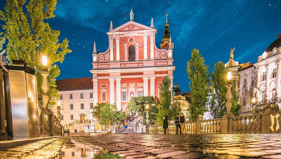 Velika roza zgradba ponoci, sredi mestnega trga, obdana z zelenjem in lucmi.