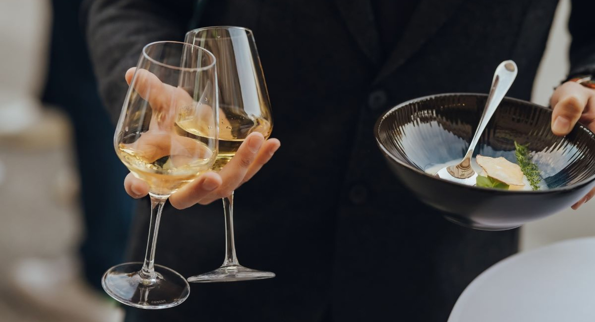 Man holding two white wine glasses and a black place with some kind of soup in his hands.