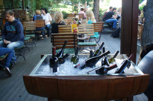 Gosti puba na zunanji terasi za lesenimi mizami. V ospredju hladilna posoda z vini, narejena iz soda.