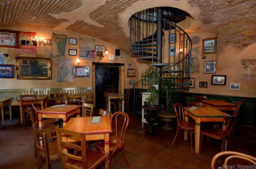 The interior of a restaurant with wooden tables and chairs. In the background, there is a circular staircase leading to an upper floor.