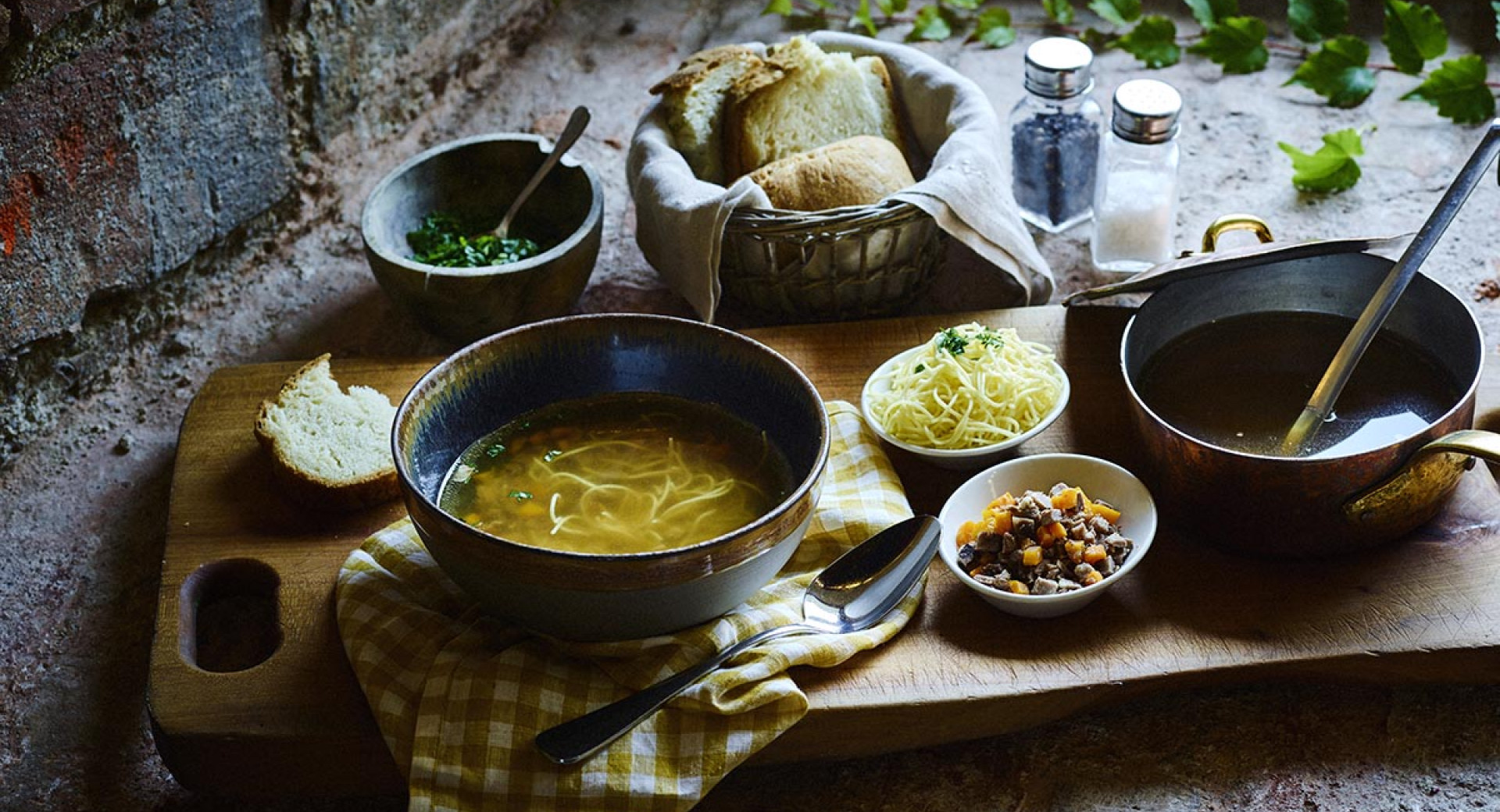 Goveja juha z rezanci, ocvirki in kruh na leseni deski. Ob deski v posodici peteršilj, kruh ter sol in poper.